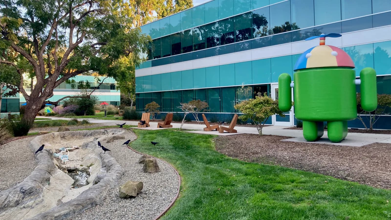 Зелёная фигура маскота Android в шапке с пропеллером возле офисного здания