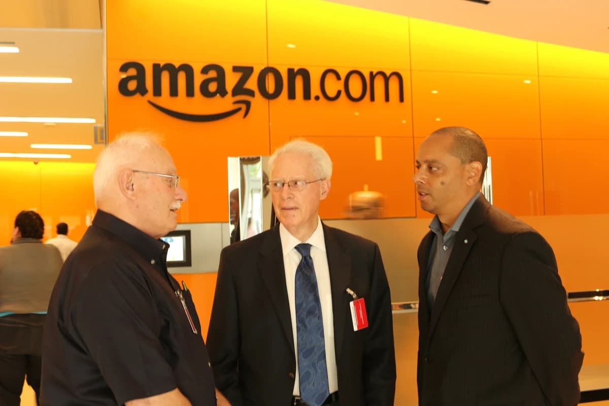 Трое мужчин беседуют в помещении на фоне крупной вывески Amazon.com
