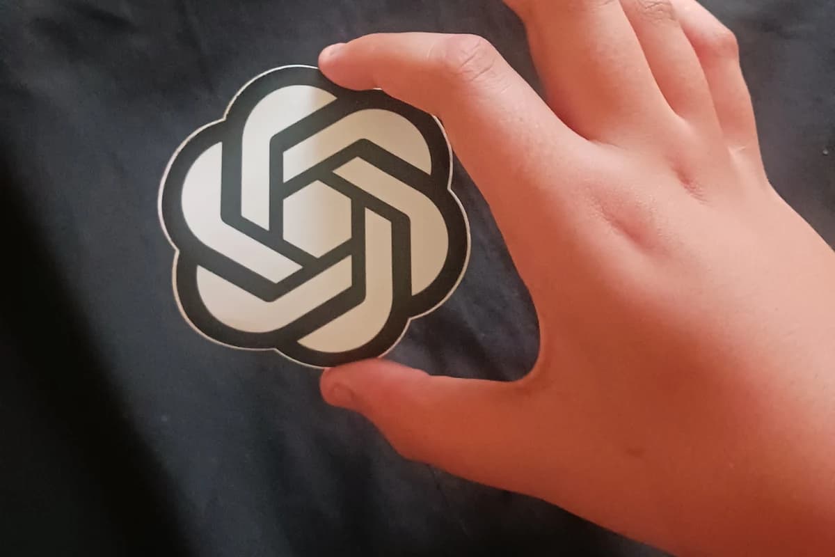 Рука держит вырезанный логотип OpenAI на тёмном фоне.