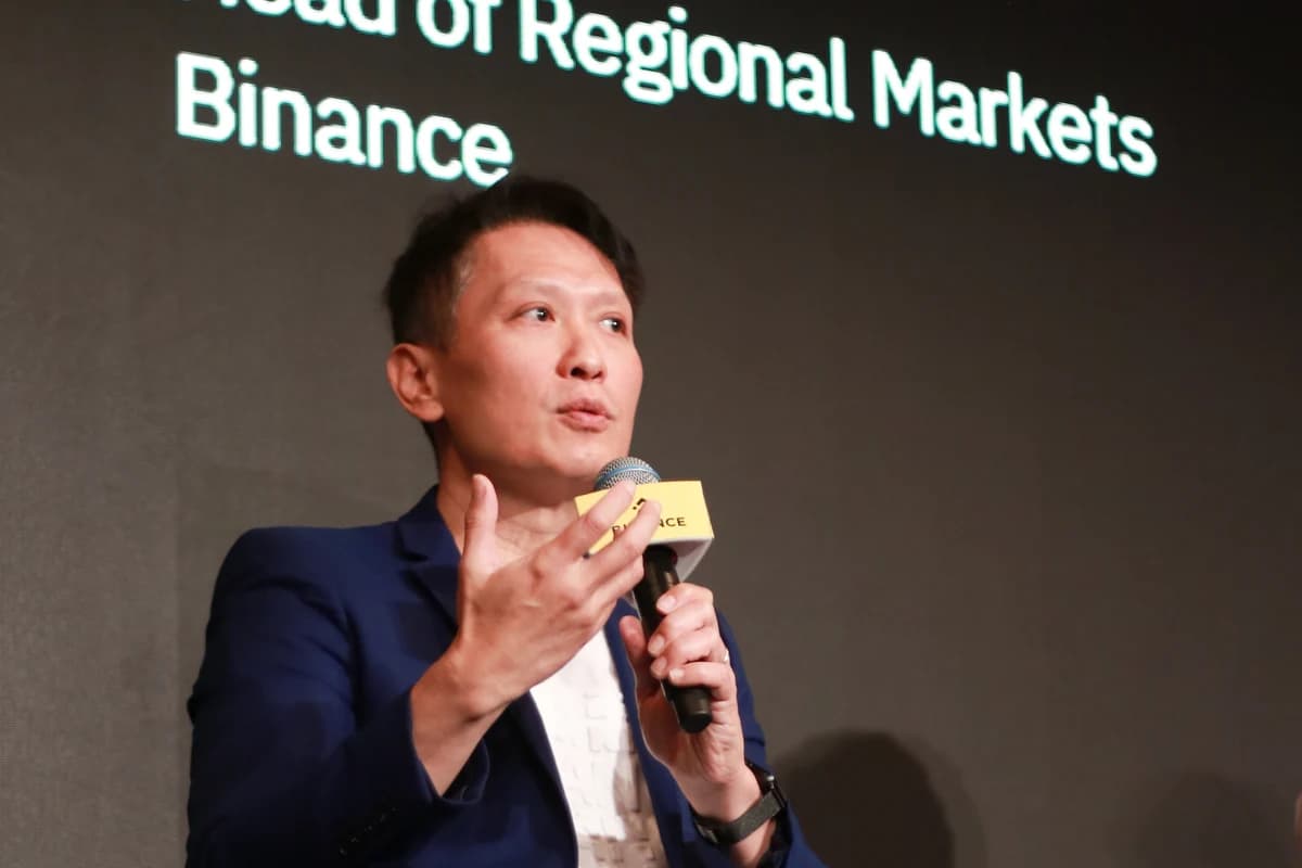 Ричард Тенг сидит на сцене с микрофоном на фоне надписи Binance